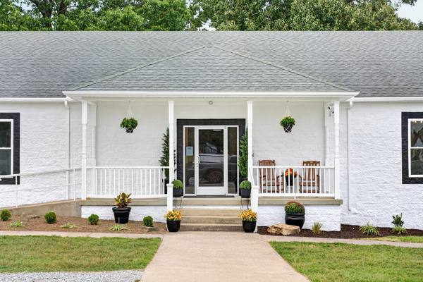 White brick building with a covered front porch, rocking chairs, hanging ferns and potted mums by the steps
