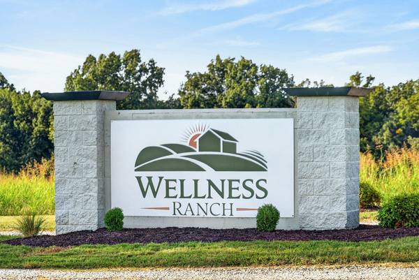 Wellness Ranch stone entrance sign with its green farm logo, shrubs and mulch bed under a blue sky