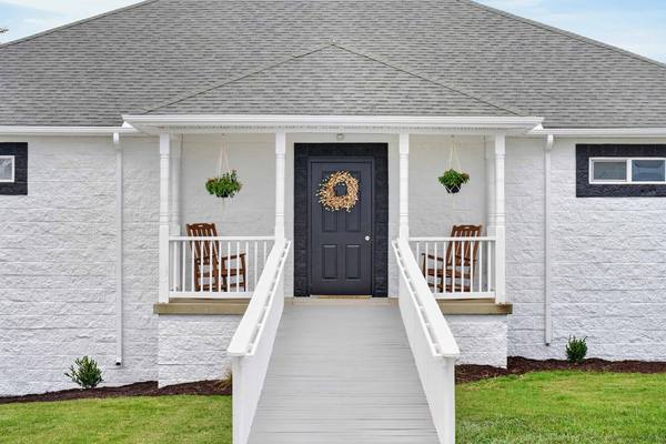 White ramp rising to a black front door with a wreath, flanked by rocking chairs and hanging plants