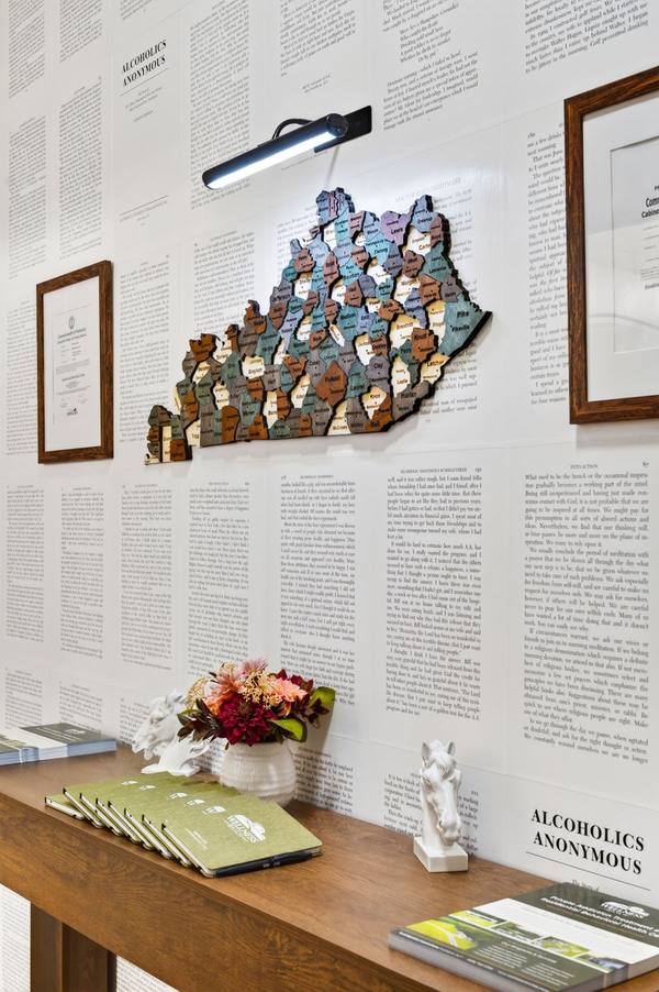 Layered wooden map of Kentucky counties on a wall papered with Alcoholics Anonymous book pages