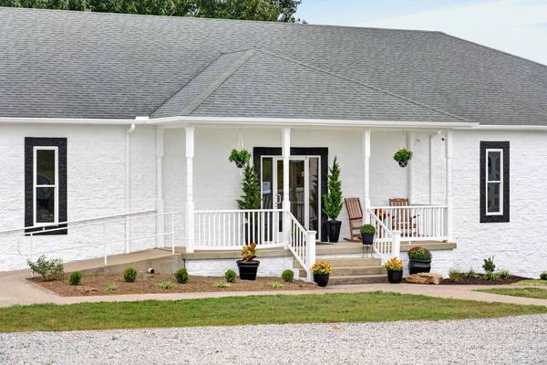 White brick building with a covered porch, rocking chairs, hanging baskets and a ramp to the door