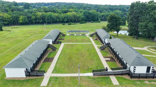 Aerial view of two long residence buildings set around a central lawn, with the pond and woods beyond