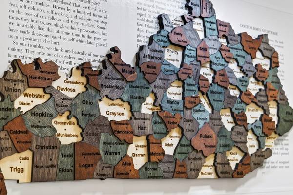 Carved wooden map of Kentucky with every county named, mounted on a wall printed with recovery text