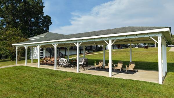 Open-air pavilion with rocking chairs and lounge seating on a concrete floor, fields beyond