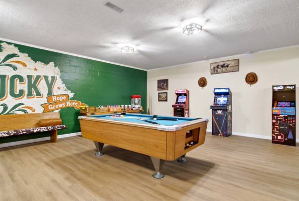Pool table with blue felt beside three lit arcade cabinets and cowboy hats hung on the wall