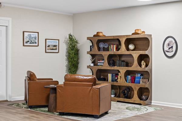Two tan leather armchairs beside a curved wooden bookshelf holding books, cowboy hats and framed landscapes